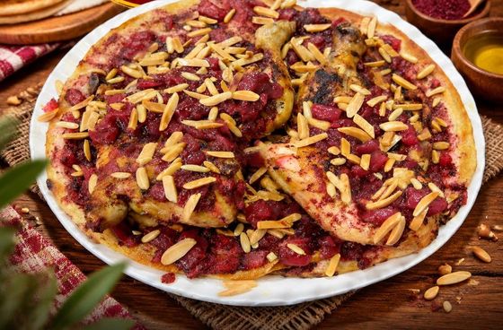 A whole mosakhan on a white plate: roast chicken on taboon bread under onions with sumac and toasted almonds